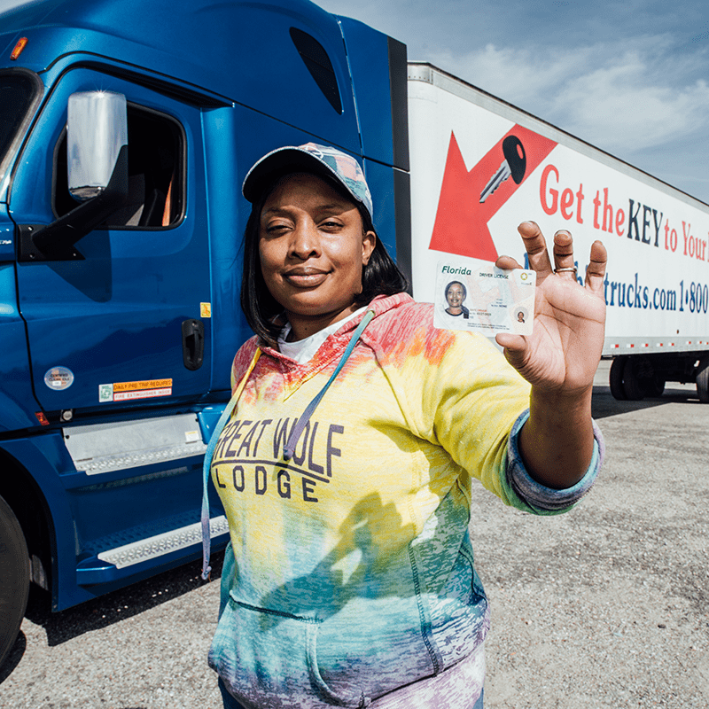 A proud truck driver stands in front of a blue semi-truck, holding up a driver's license.