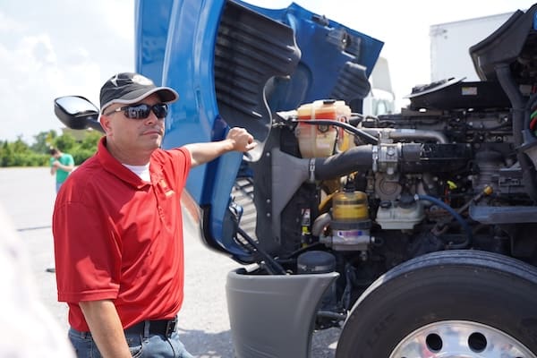 An instructor stands next to an open hood of a commercial truck following all CDL requirements by state