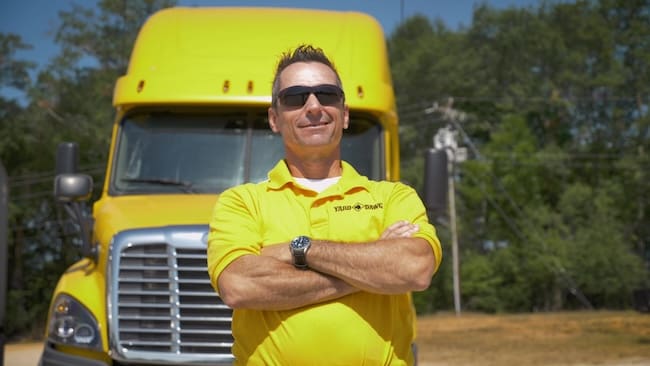 A driver stands with arms folded in front of a yellow commercial truck