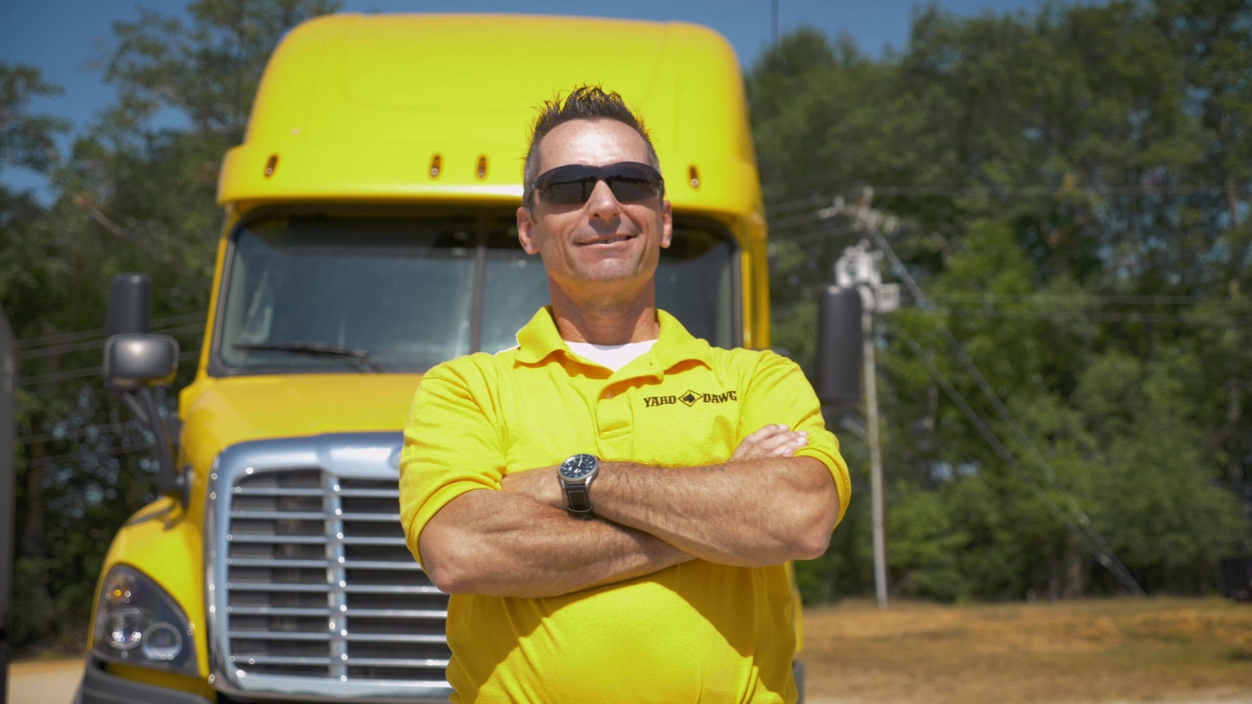 A man in a yellow shirt stands proudly in front of a yellow truck after passing his CDL classes.
