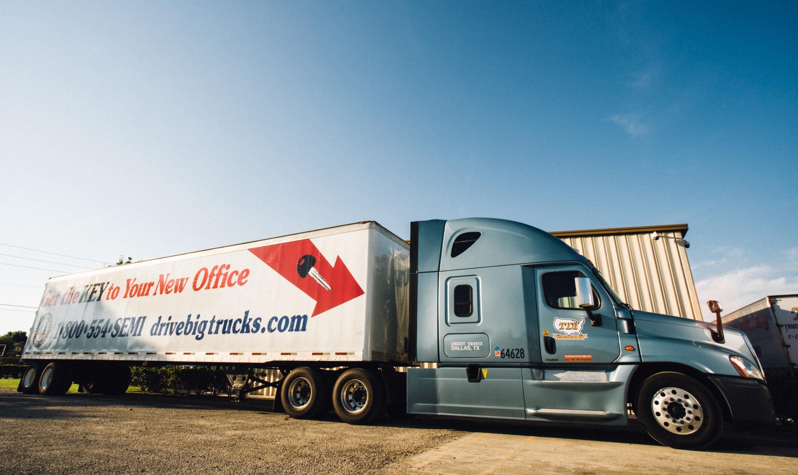 A large white-and-blue semi-truck parked outside on a sunny afternoon. The side of the truck reads "Get the KEY to Your New Office: 1-800-554-SEMI drivebigtrucks.com." You'll need to get your CDL permit first before your CDL license to drive trucks like these.